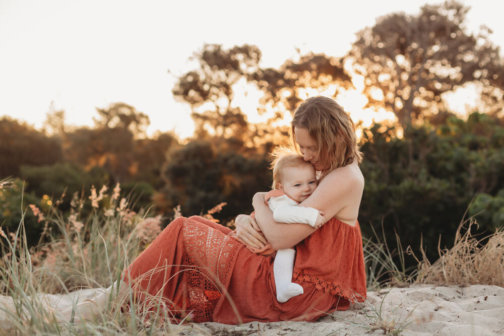 Mum holds baby girl in a grassy field while the sunsets behind them.