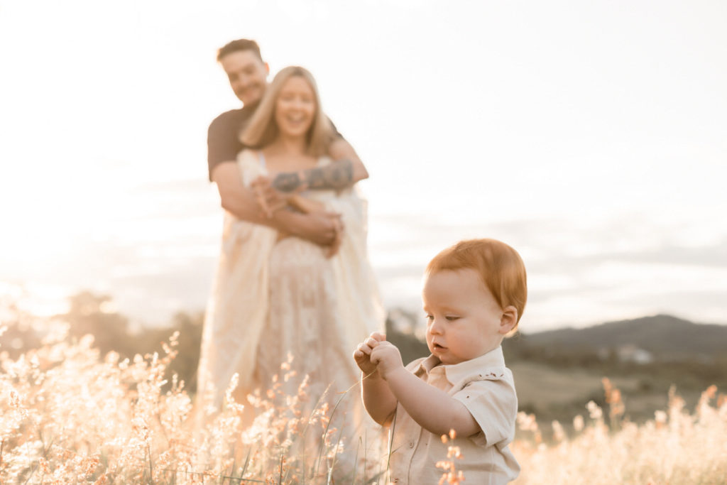 Husband holding pregnant Mama in sheer lace dress, while they look at their toddler in the foreground. A Golden Hour Shoot