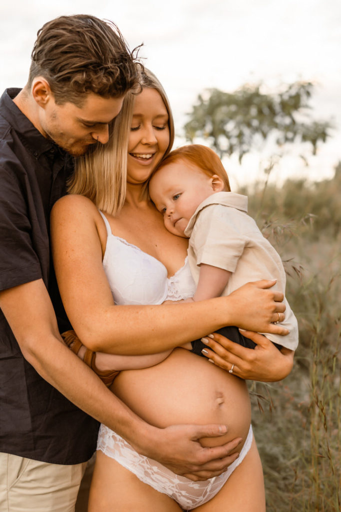 Husband holding pregnant Mama in sheer lace dress, while they look at their toddler in in mamas arms. A Golden Hour Shoot