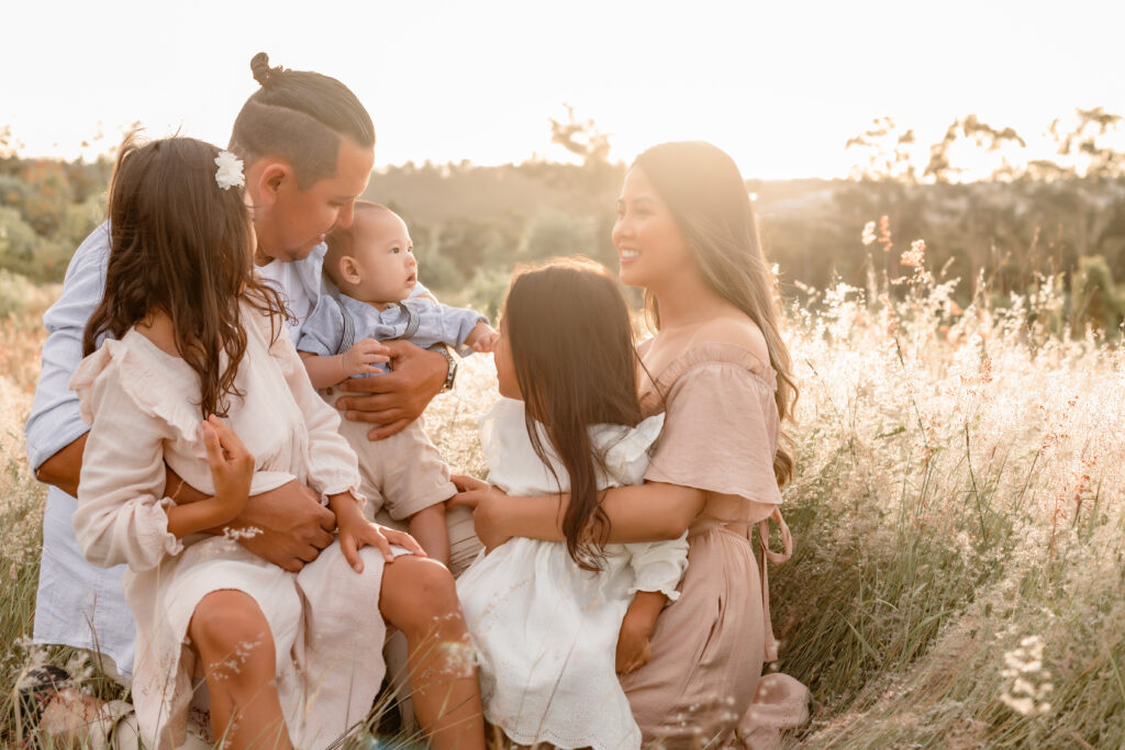 A famly of five are seated, cuddling in a wheat field with a sunset behind them.
