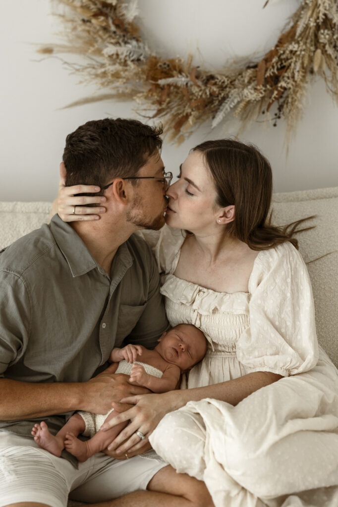 A husband and wife with thier newborn, lounging on a white couch with a dried plant wreath behind them