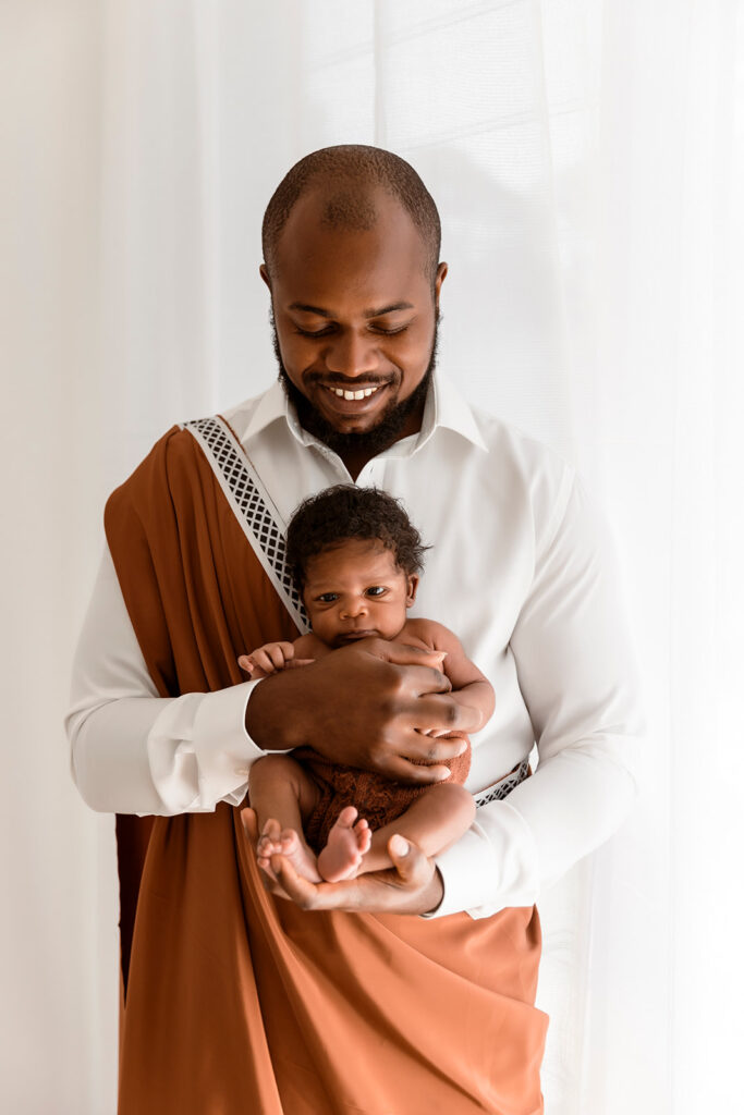 Dad holding bub, facing the camera, while looking down at him. Dad and bub are wearing cultural attire.